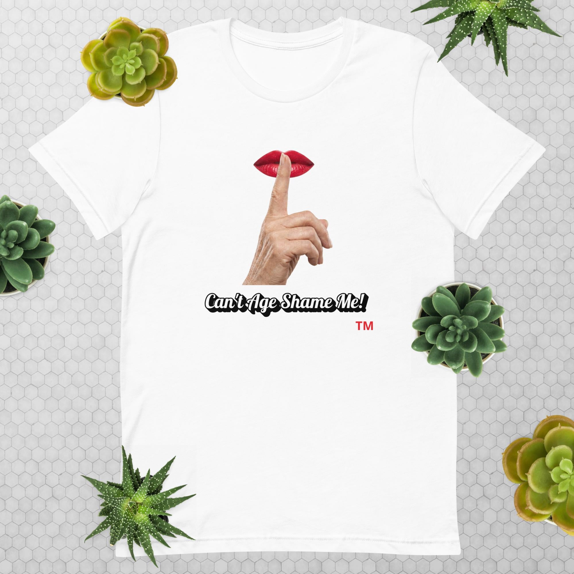 White t-shirt with graphic design of finger on mouth and text 'CAN'T AGE SHAME ME!' text logo on a light gray background with succulents.