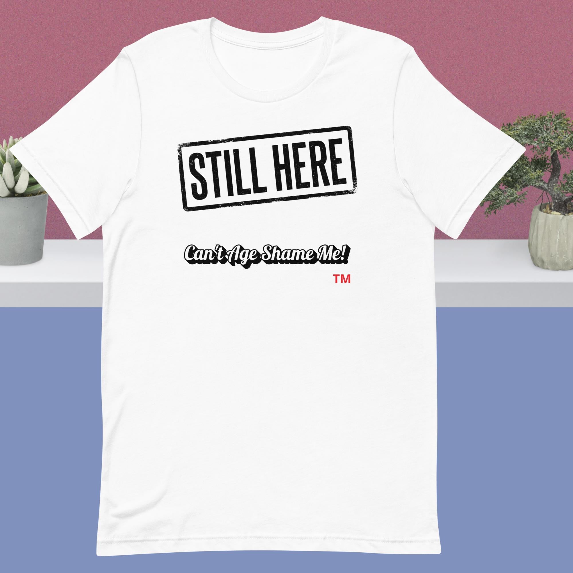 White t-shirt with 'STILL HERE' text on a white surface with plants in the background