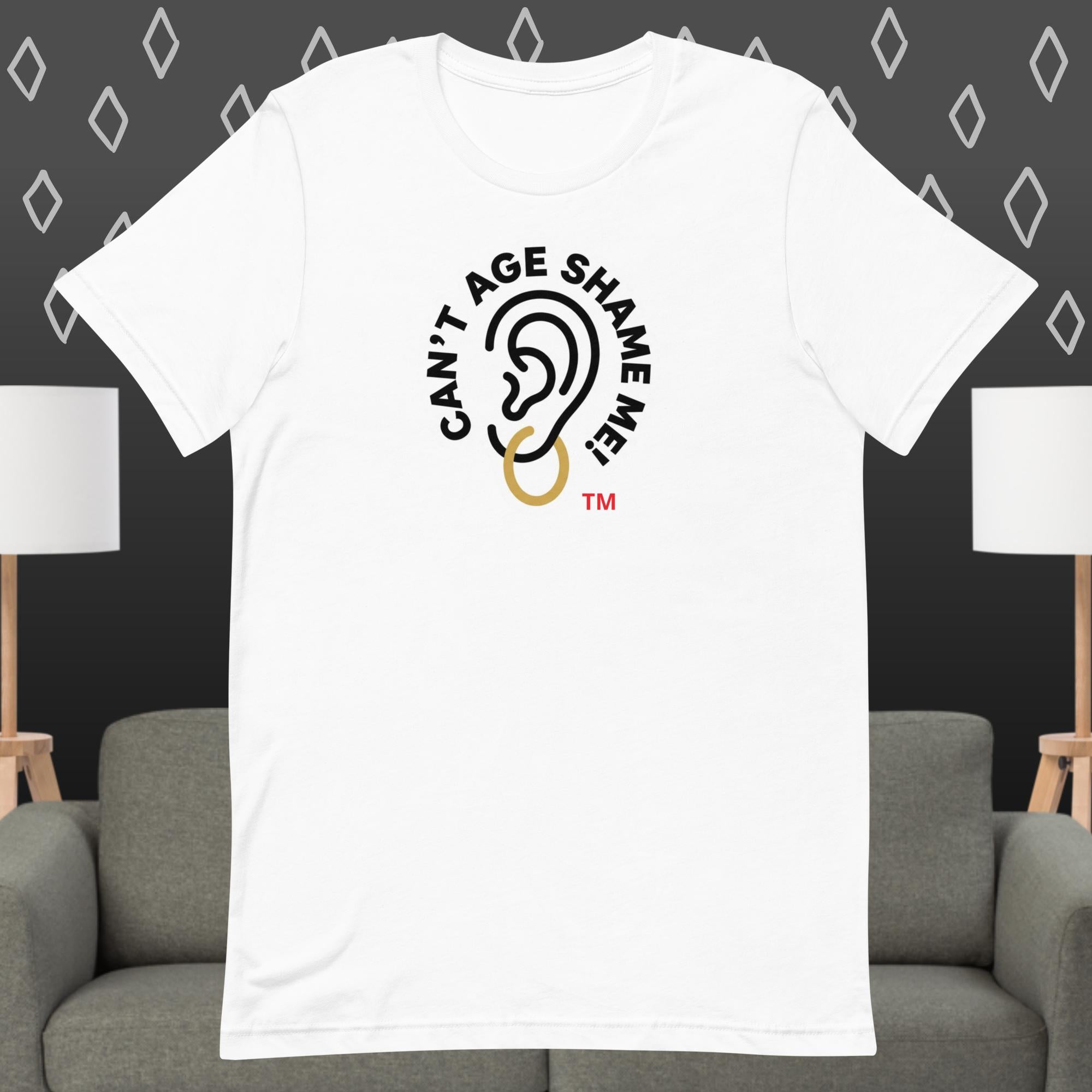 White t-shirt with 'Can't Age Shame Me' graphic on a gray couch and lamp background