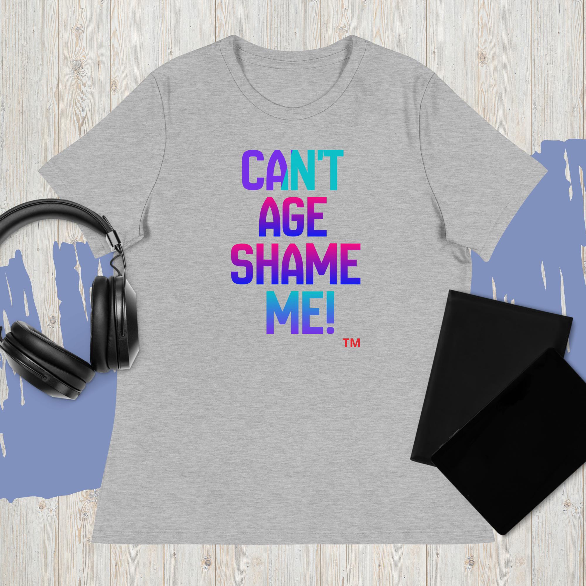 Gray t-shirt with colorful text 'CAN'T AGE SHAME ME!' on a wooden surface with headphones and a black mat.