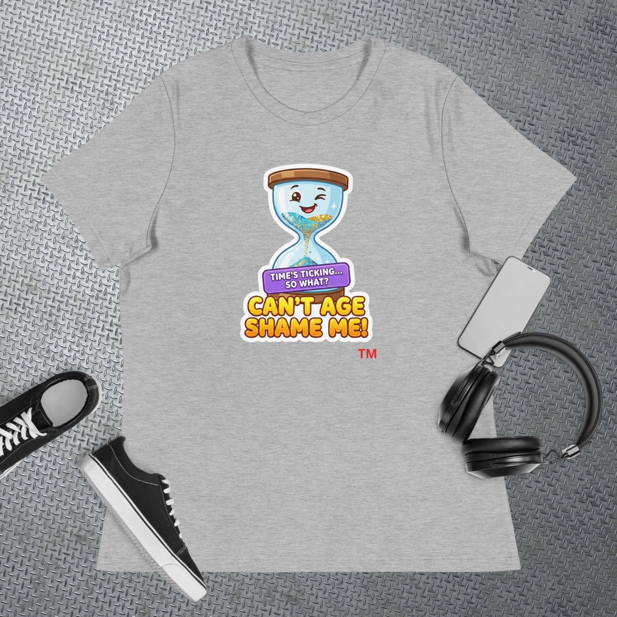 Light gray t-shirt with a cartoon hourglass graphic design and text 'Can't Age Shame Me' graphic on a textured gray surface with a sneaker and headphones