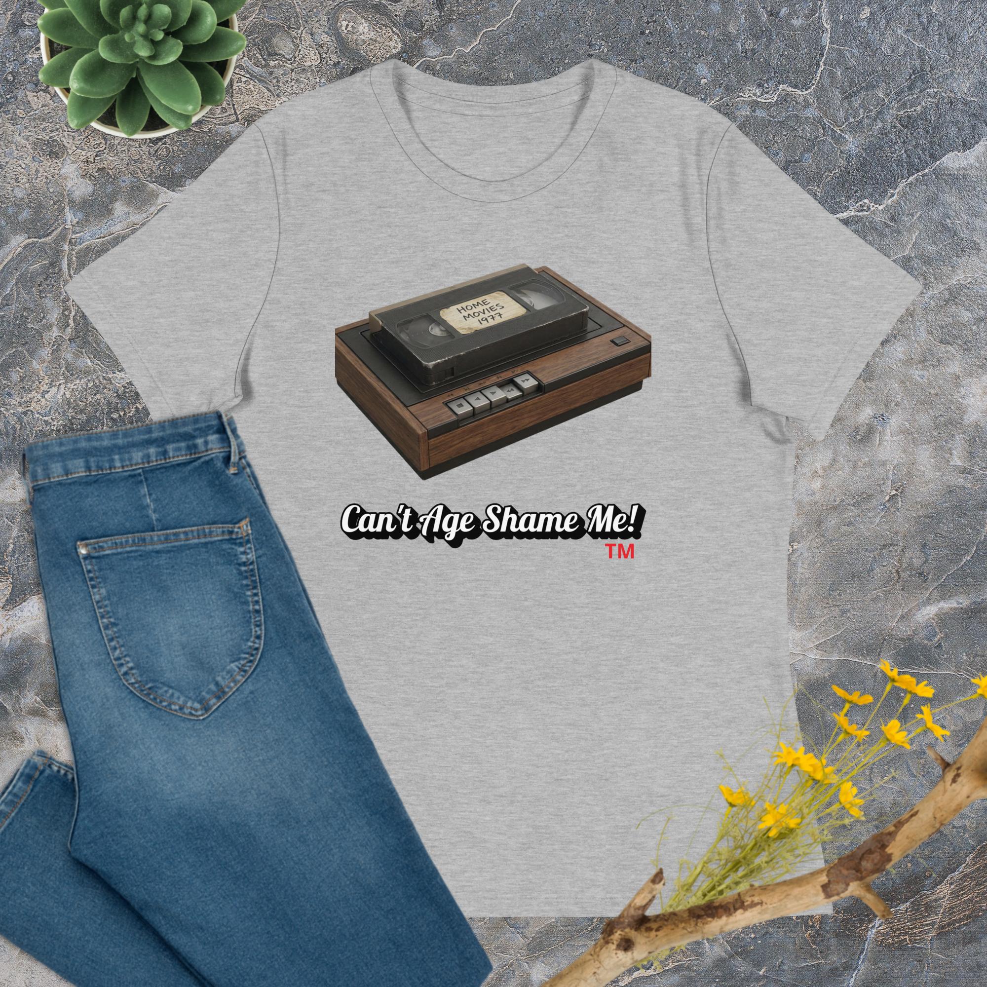 athletic heather t-shirt with a vintage VHS tape graphic and 'Can't Age Shame Me!' text, paired with blue jeans on a stone surface.