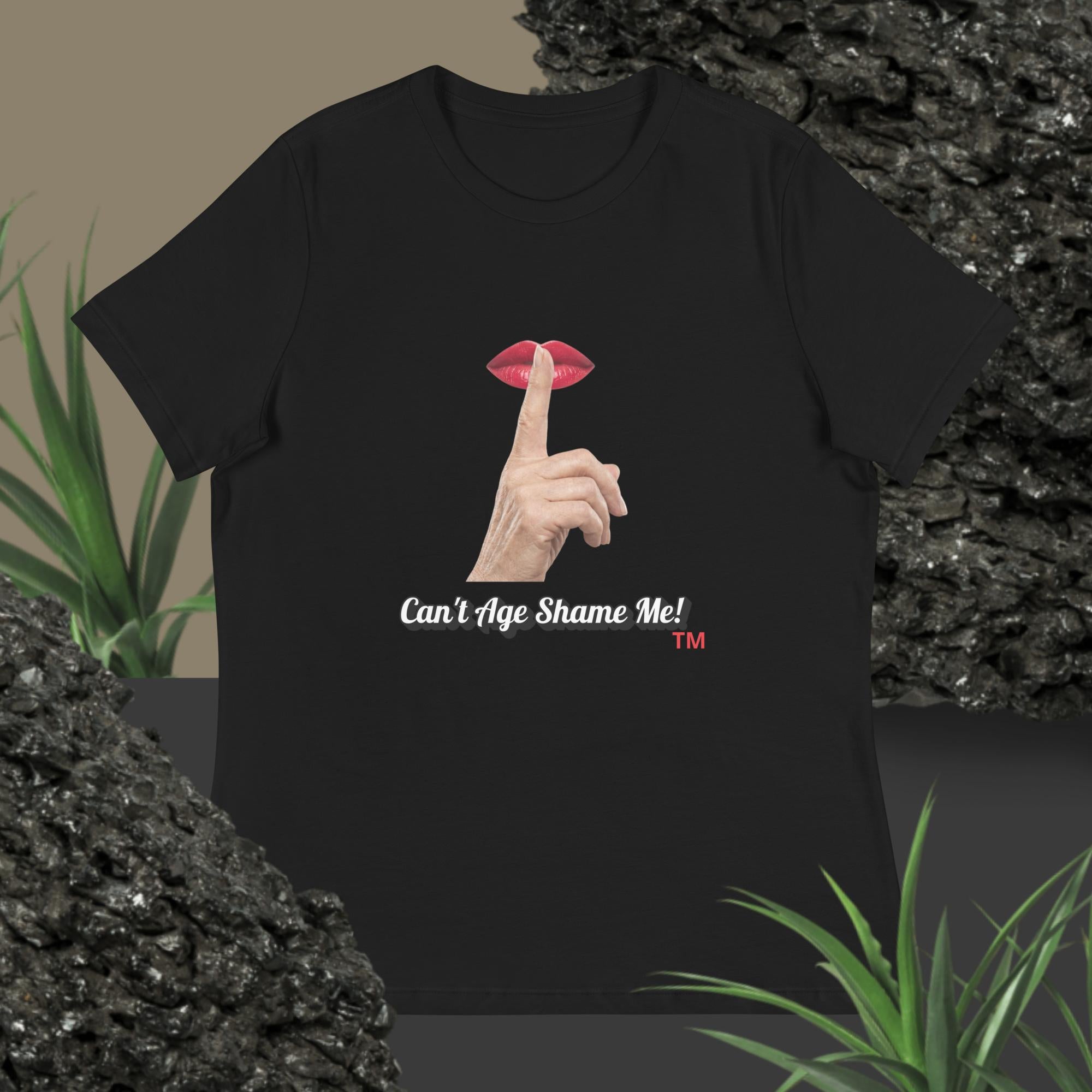 Black t-shirt with graphic design of finger on mouth and text 'Can't Age Shame Me' on a textured surface with plants.