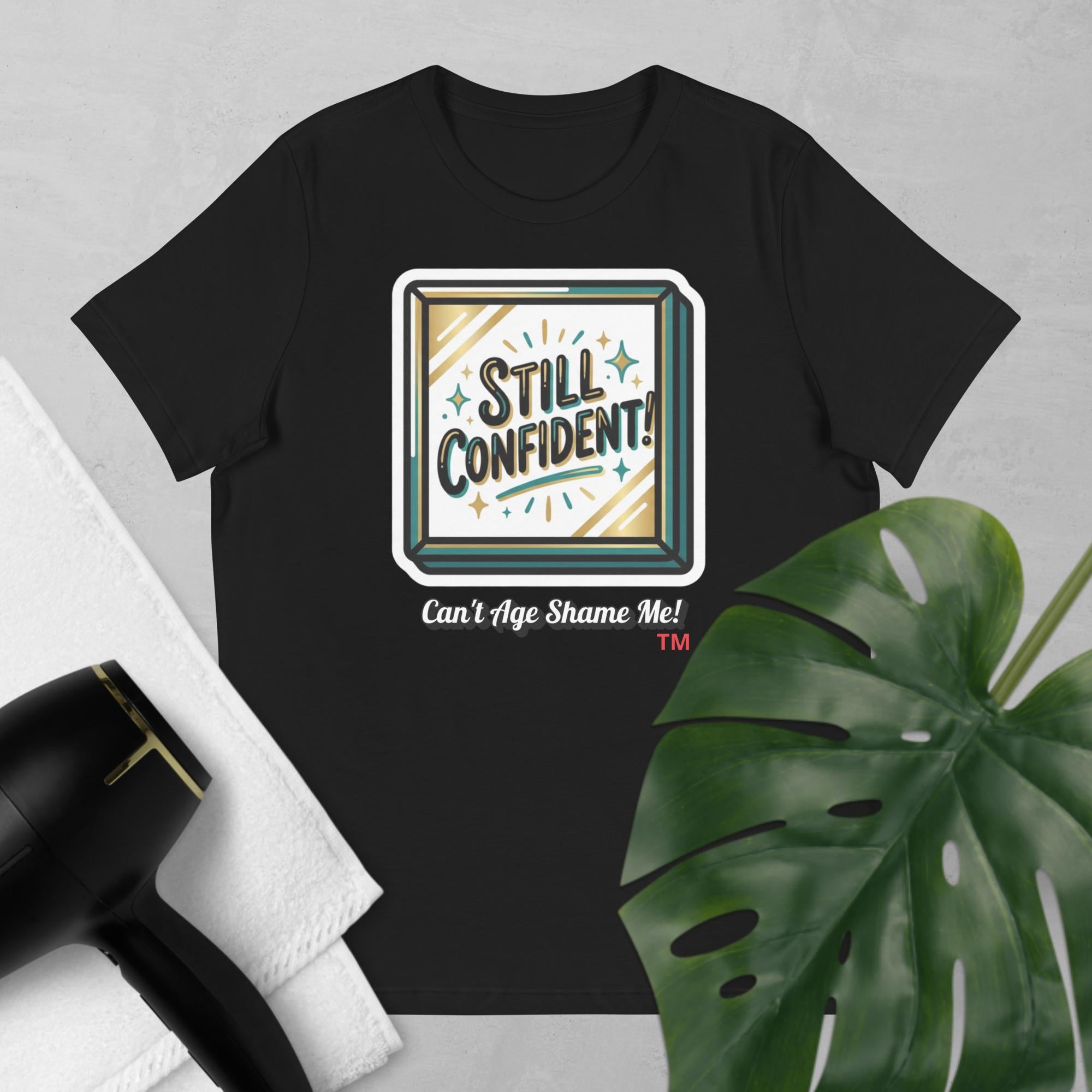 Black t-shirt with 'Still Confident! Can't Age Shame Me!' design on a gray background with a plant and hair dryer.