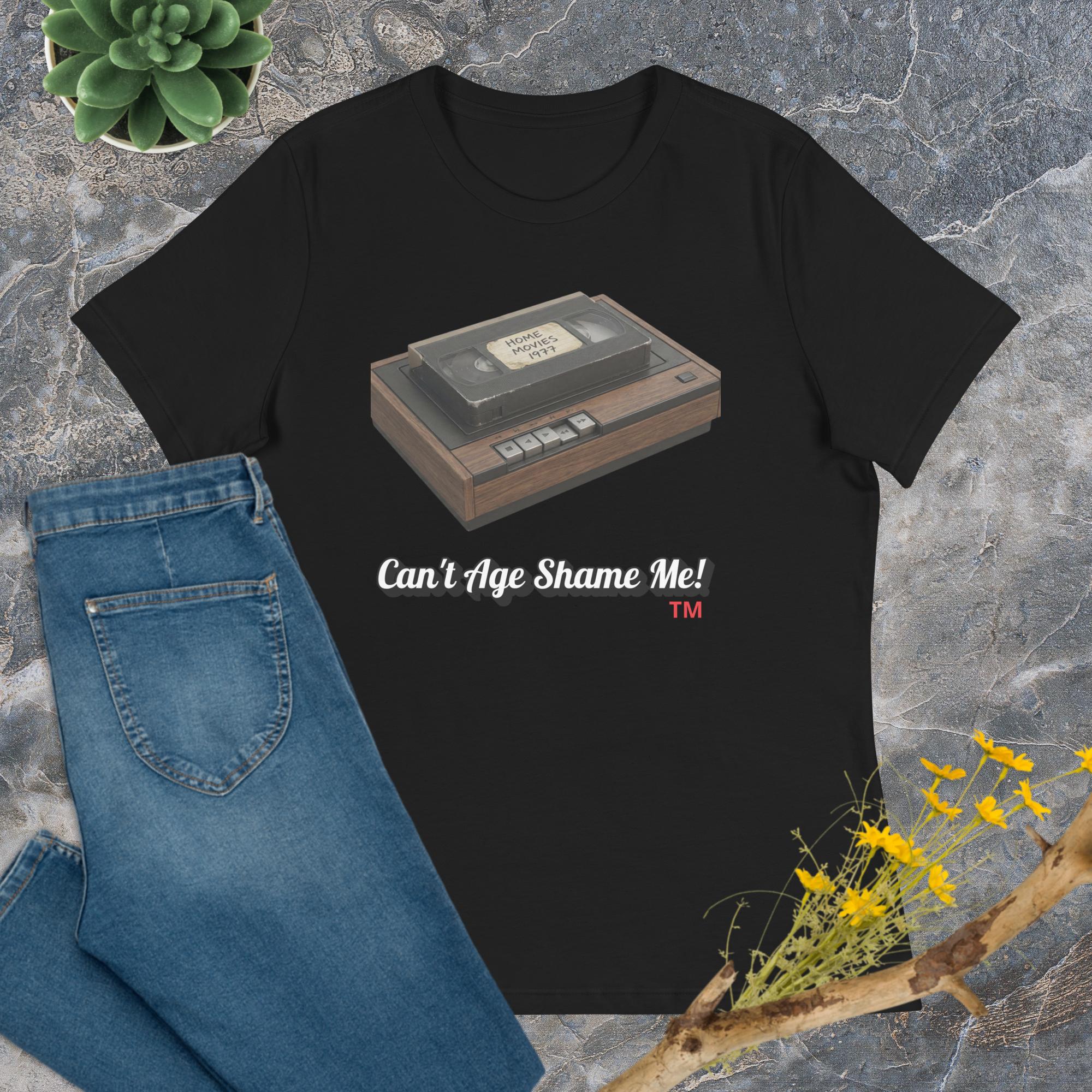 Black t-shirt with a cassette tape graphic and text 'Can't Age Shame Me!' on a textured surface with jeans and plants.