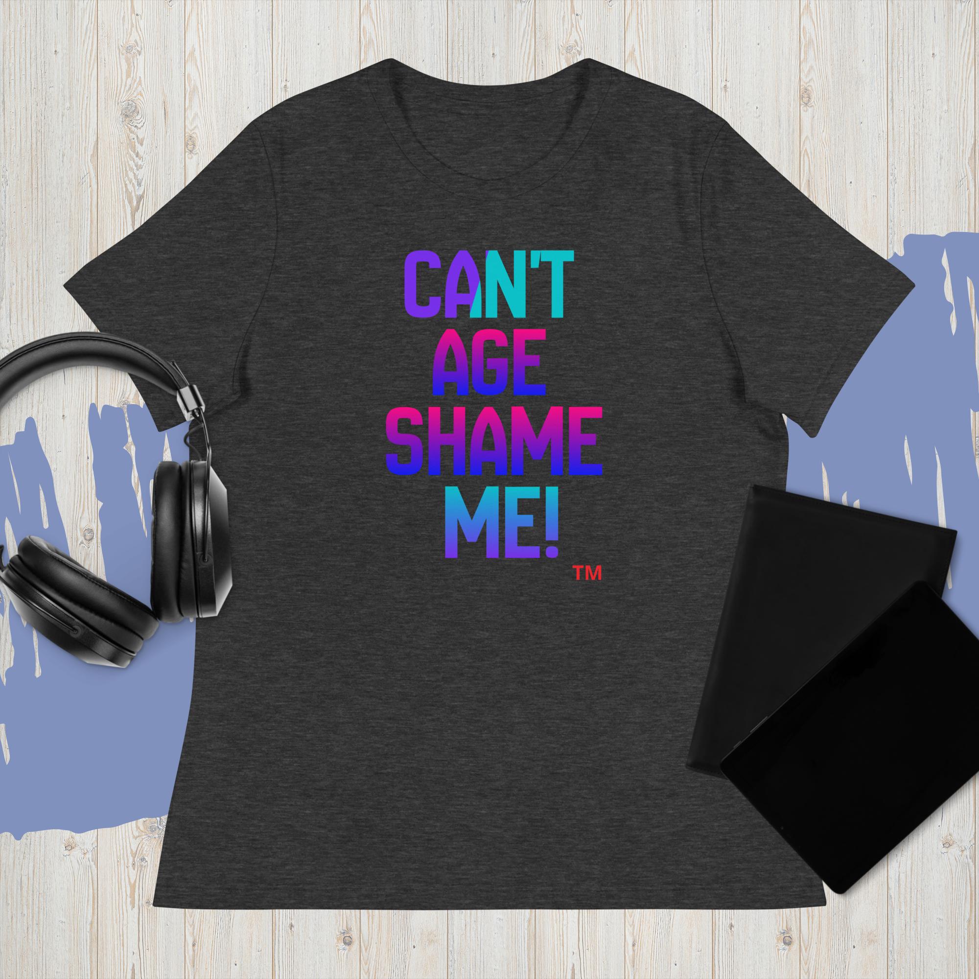 Dark gray t-shirt with colorful text 'CAN'T AGE SHAME ME!' on a wooden surface with headphones and a black pouch.