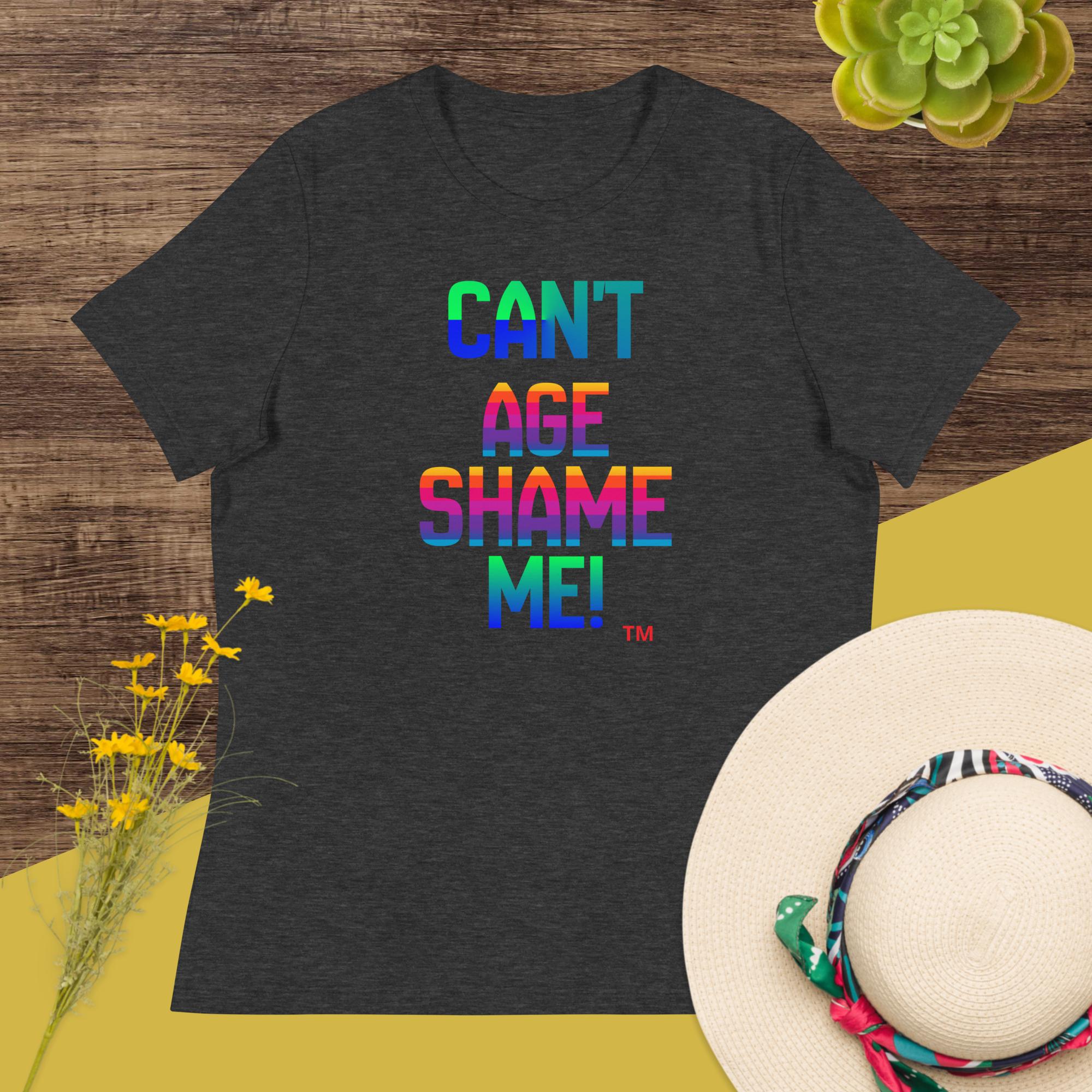 Dark gray t-shirt with colorful text 'CAN'T AGE SHAME ME!' on a wooden surface with flowers and a hat.