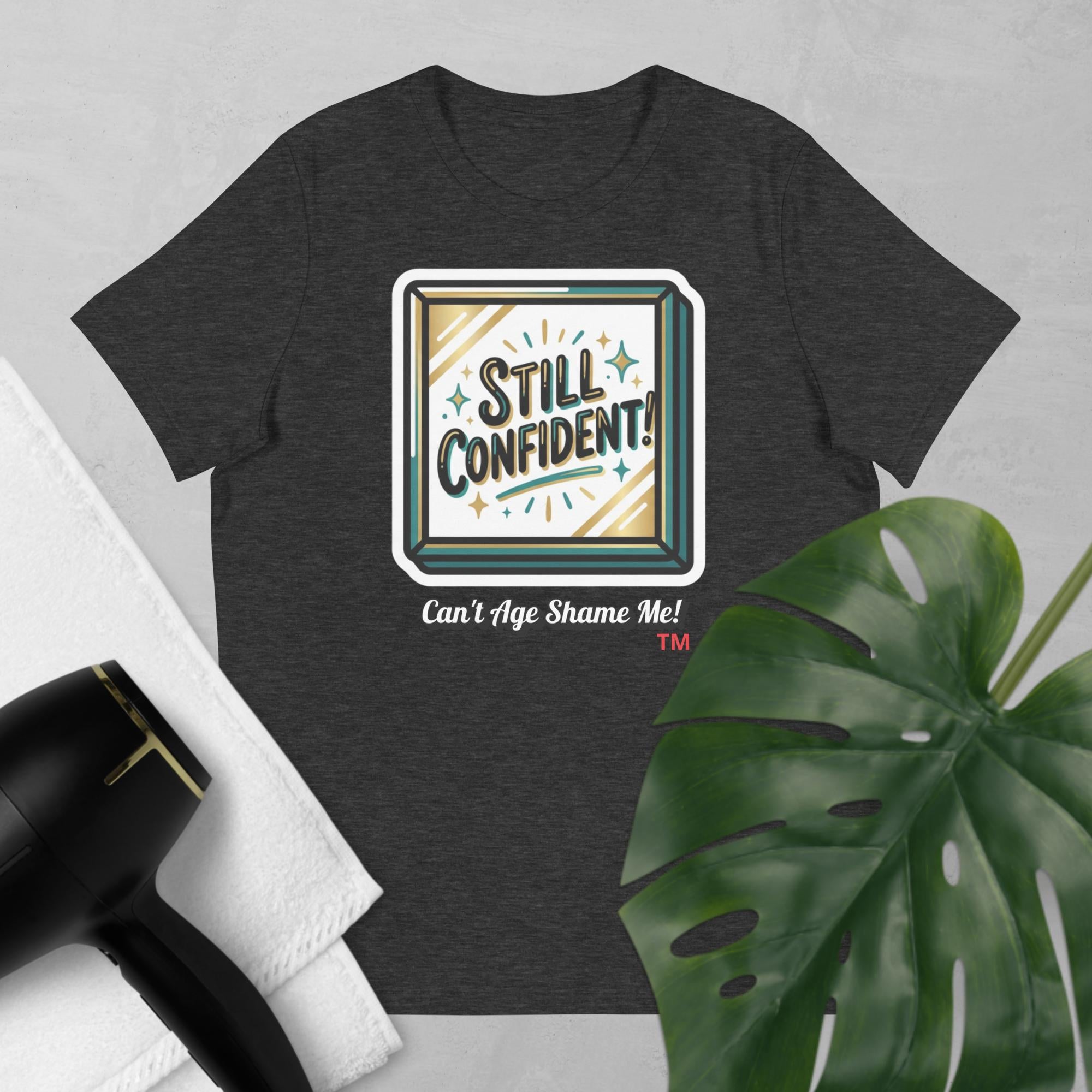 Dark Gray t-shirt with 'Still Confident! Can't Age Shame Me!' graphic on a light gray background with a plant and hair dryer.