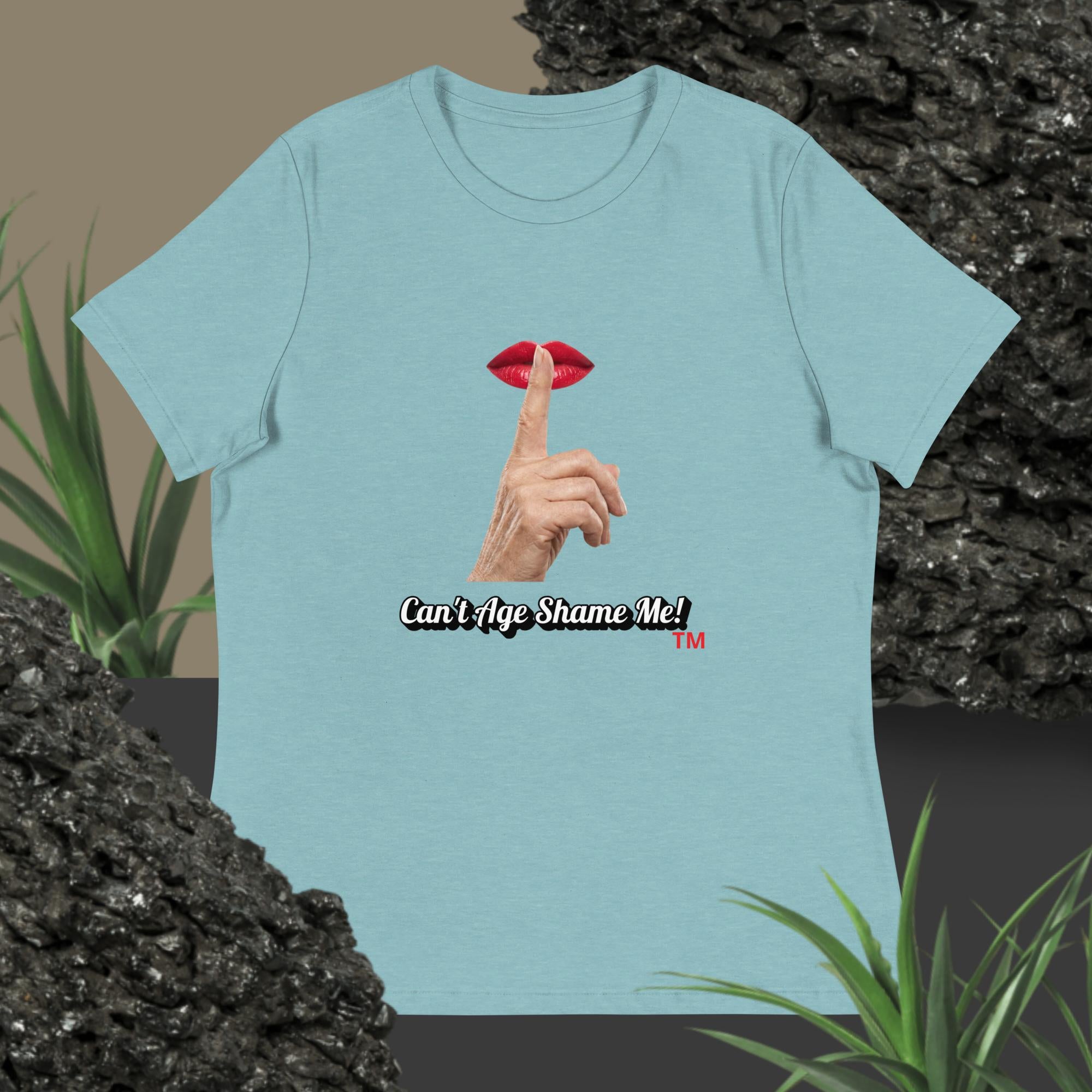 Blue Lagoon t-shirt with graphic design of finger on mouth and text 'Can't Age Shame Me' on a textured surface with plants.
