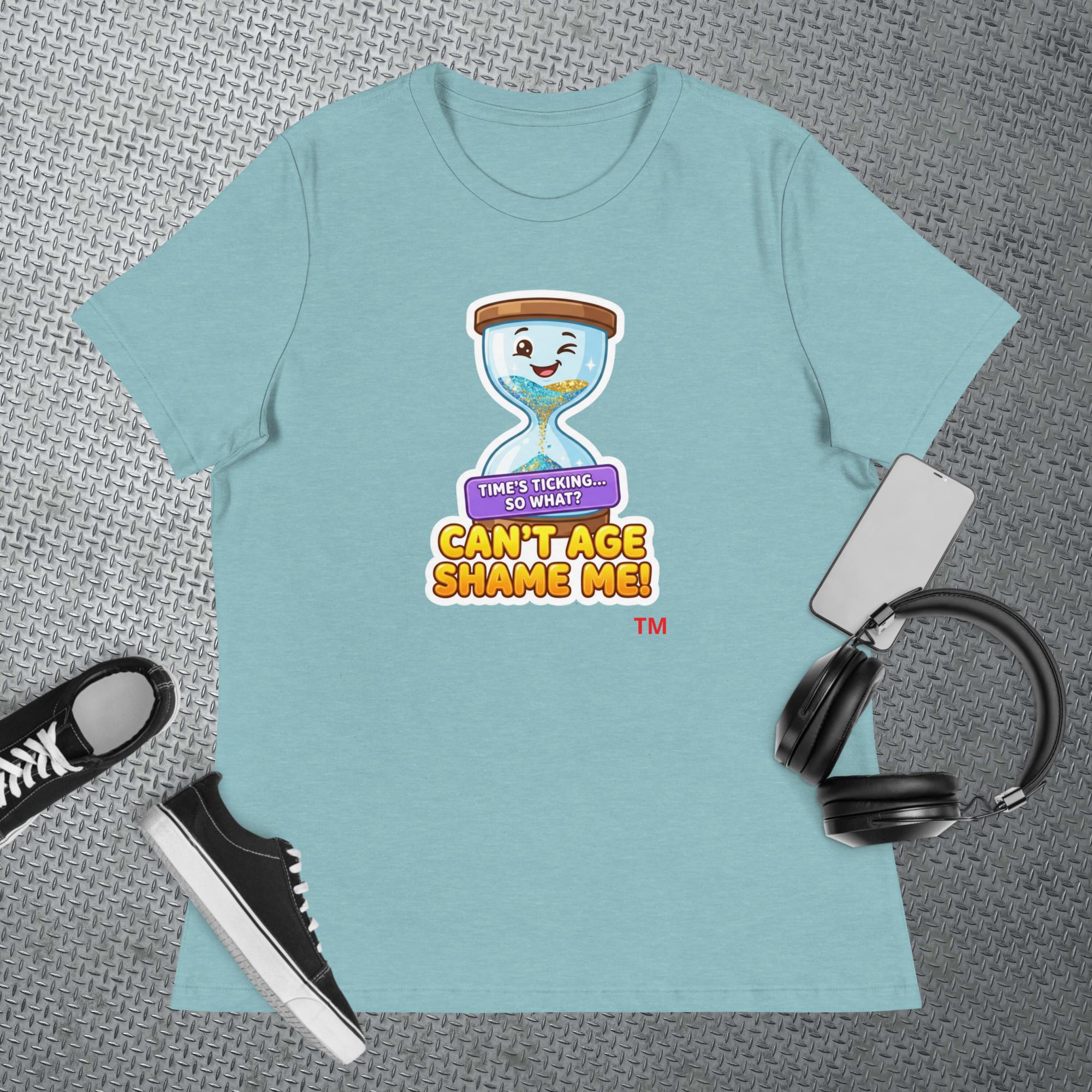 Light blue t-shirt with a cartoon hourglass graphic design and text 'Can't Age Shame Me' graphic on a textured gray surface with a sneaker and headphones