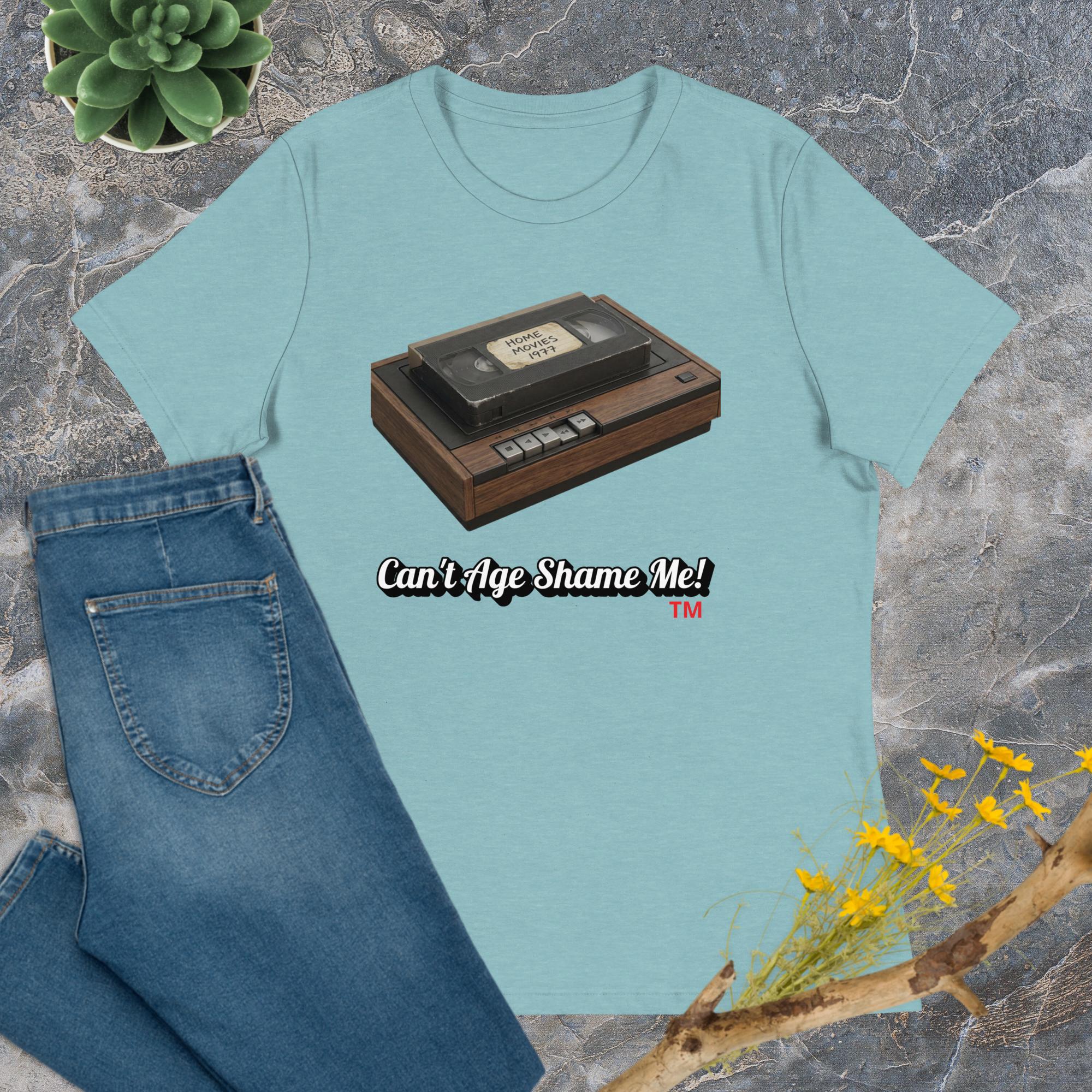 Light blue t-shirt with a cassette tape graphic and 'Can't Age Shame Me!' text on a stone surface with jeans and flowers.