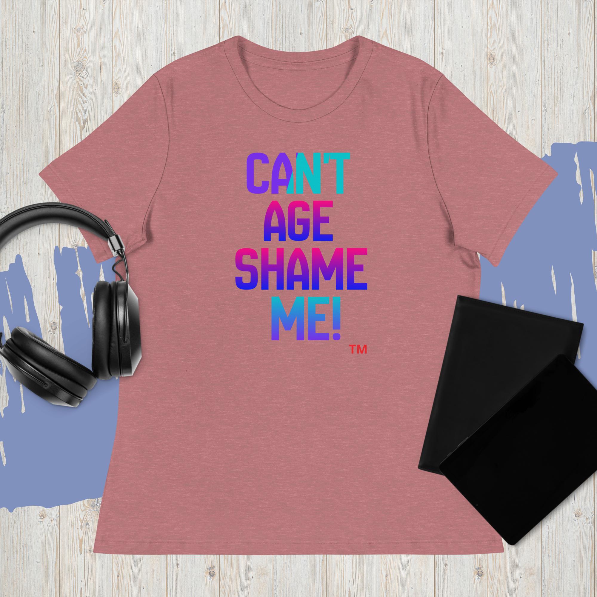 Pink t-shirt with colorful text 'CAN'T AGE SHAME ME!' on a wooden surface with headphones and a black pouch.