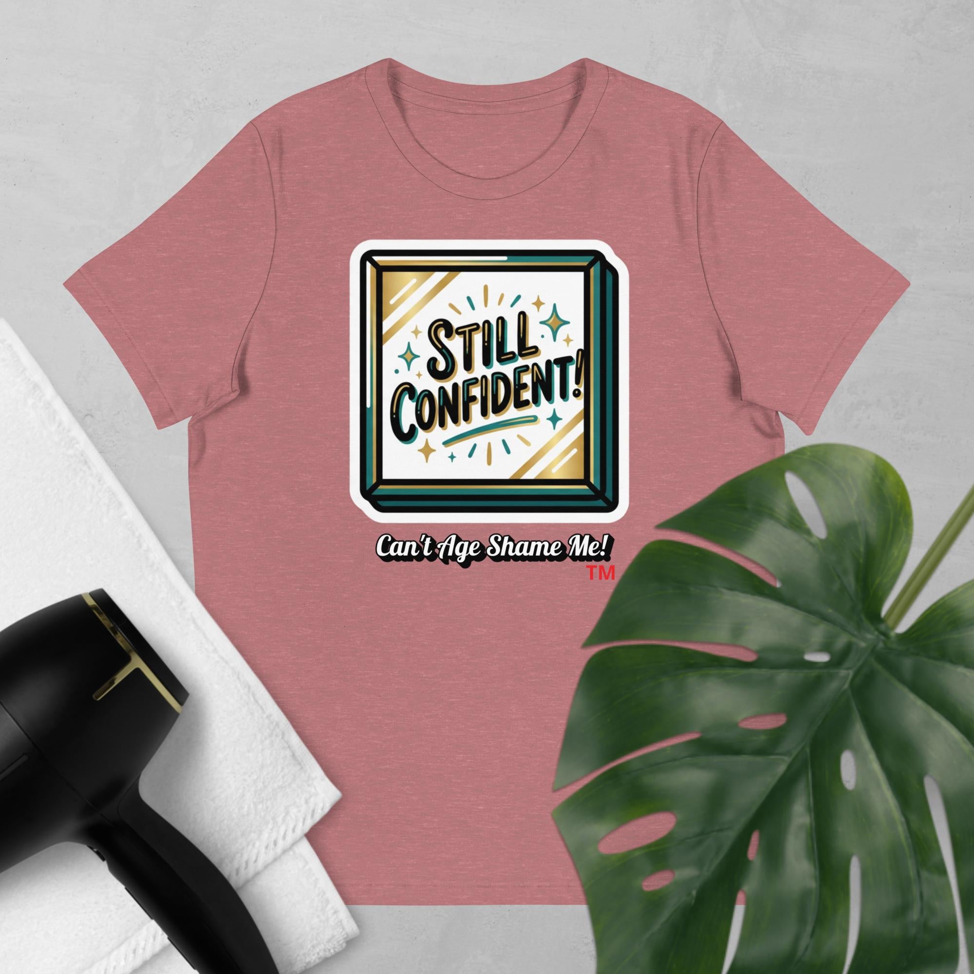 Mauve t-shirt with 'Still Confident! Can't Age Shame Me!' graphic on a light gray background with a plant and hair dryer.