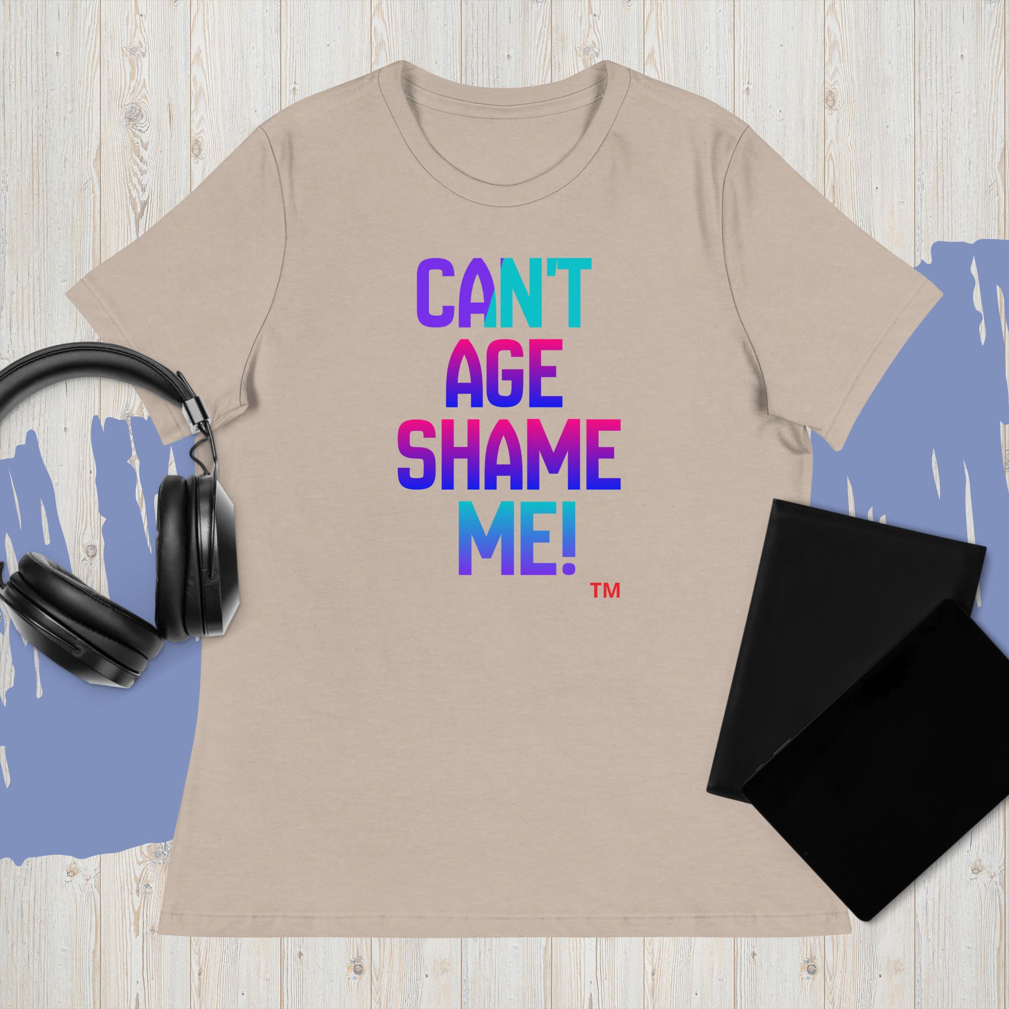 Beige t-shirt with colorful text 'CAN'T AGE SHAME ME!' on a wooden surface with headphones and a black cloth.