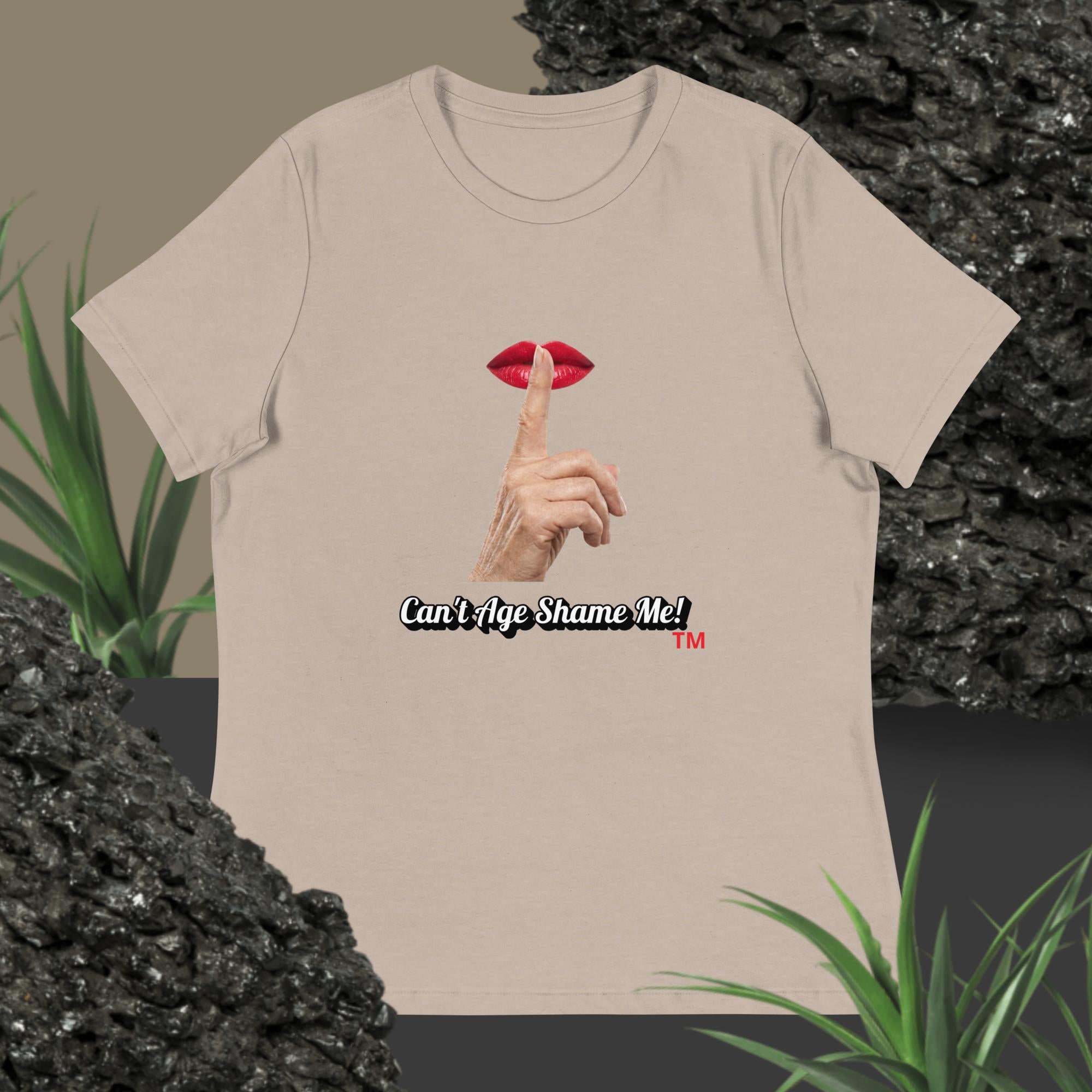 Heather Stone Pink t-shirt with graphic design of finger on mouth and text 'Can't Age Shame Me' on a textured surface with plants.