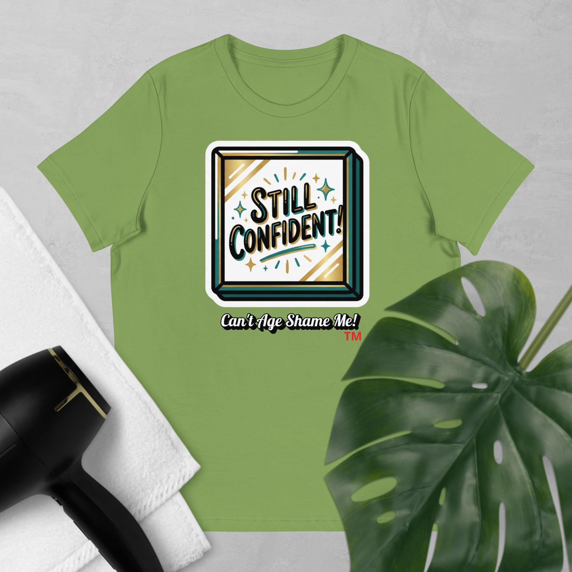 Light Green t-shirt with 'Still Confident! Can't Age Shame Me!' graphic on a light gray background with a plant and hair dryer.