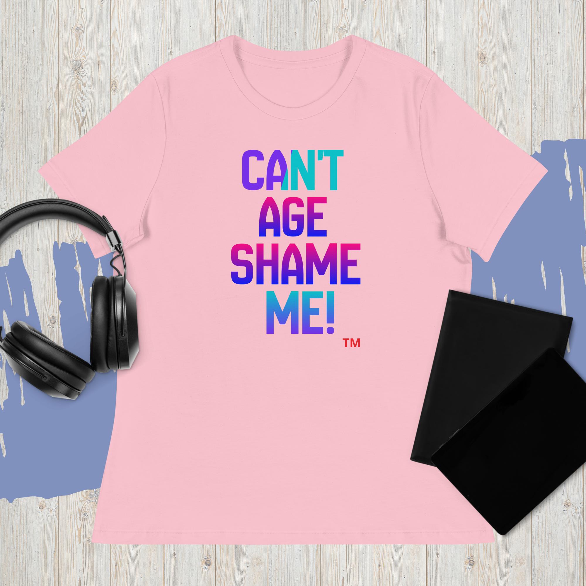 Pink t-shirt with colorful text 'CAN'T AGE SHAME ME!' on a wooden surface with headphones and a black pouch.