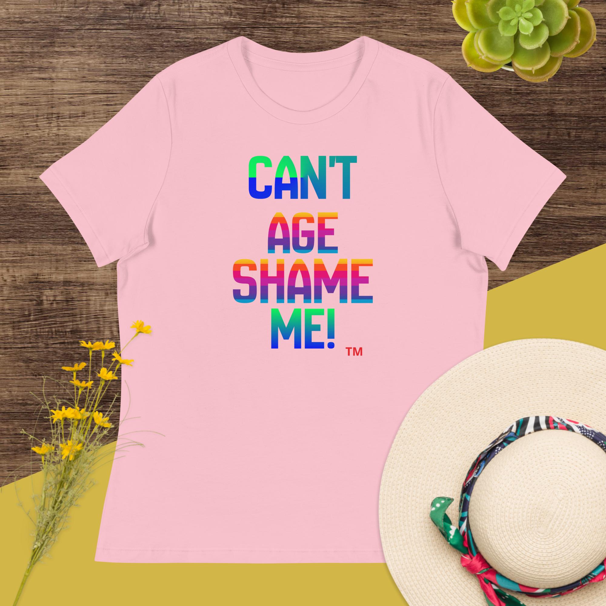 Pink t-shirt with colorful text 'CAN'T AGE SHAME ME!' on a wooden surface with flowers and a hat.
