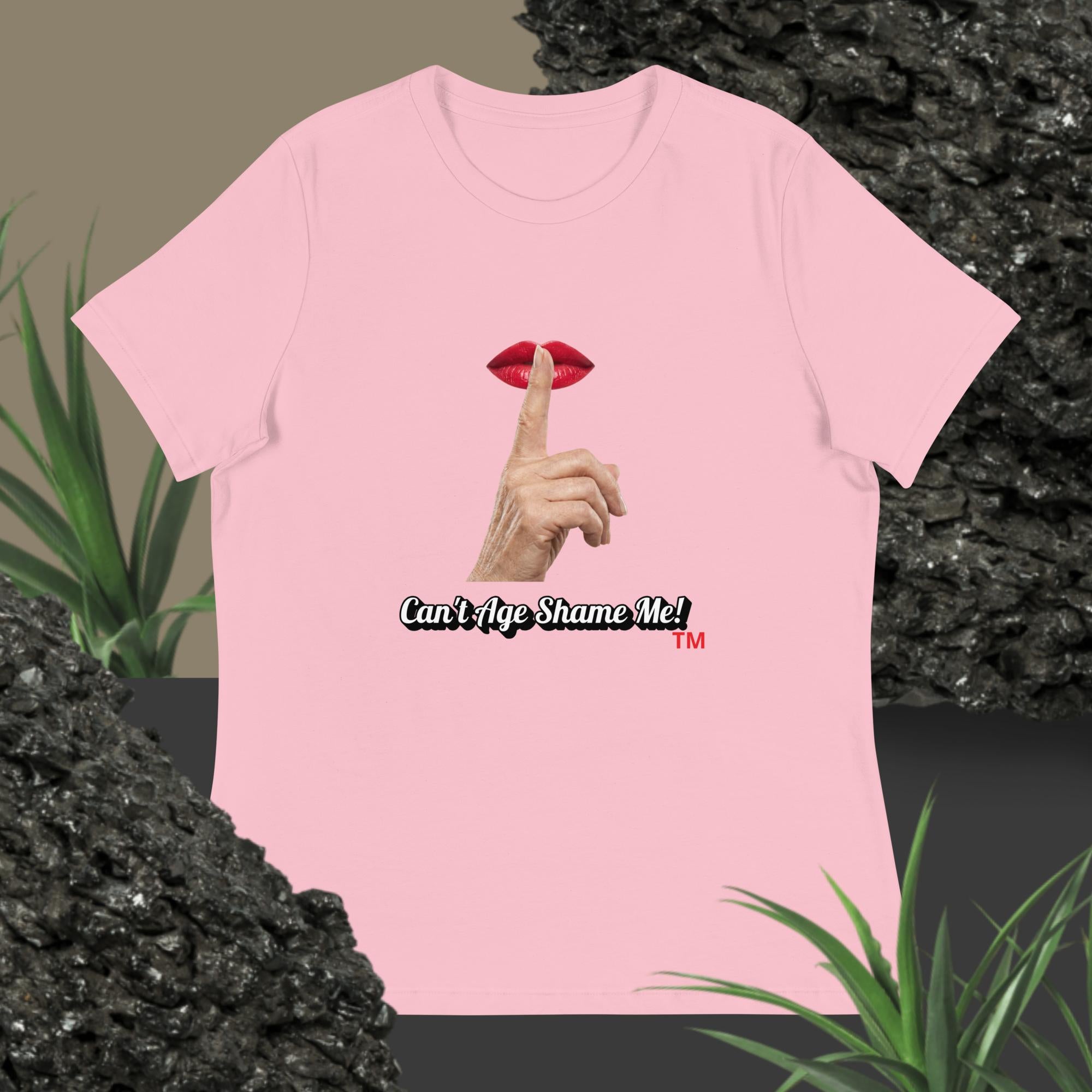 Pink t-shirt with graphic design of finger on mouth and text 'Can't Age Shame Me' on a textured surface with plants.