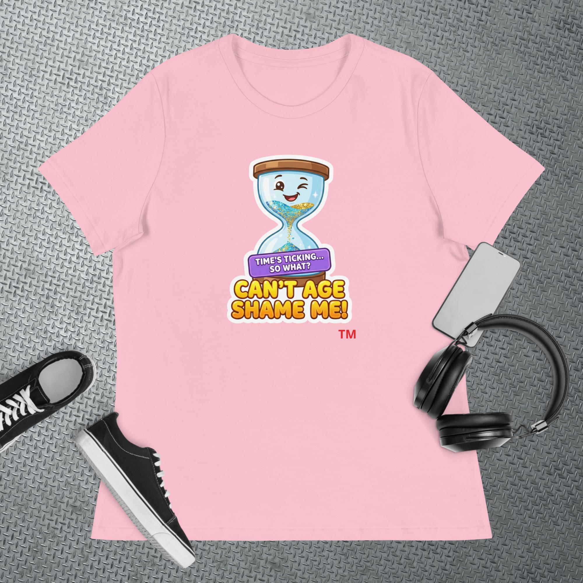 Pink t-shirt with a cartoon hourglass graphic design and text 'Can't Age Shame Me' graphic on a textured gray surface with a sneaker and headphones