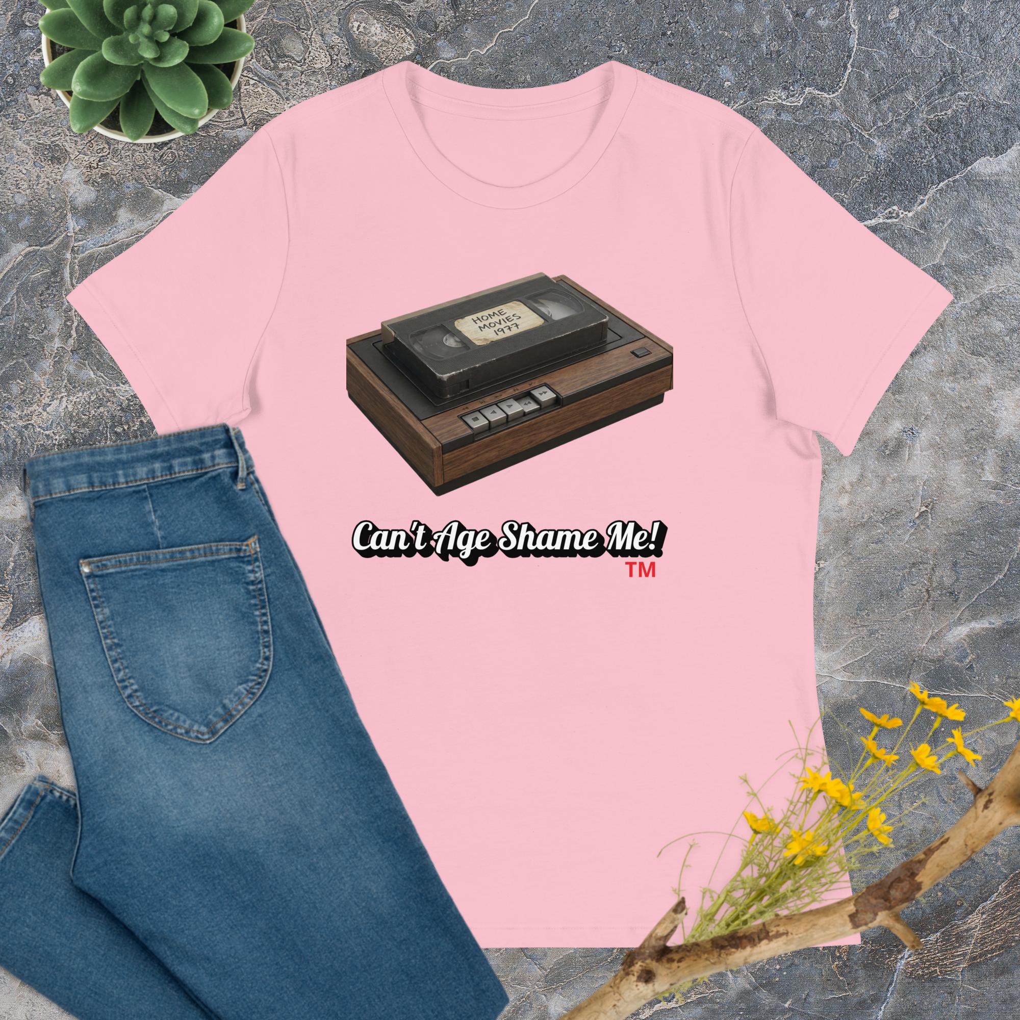 Pink t-shirt with VHS tape graphic and text 'Can't Age Shame Me!' on a textured surface with jeans and plants.