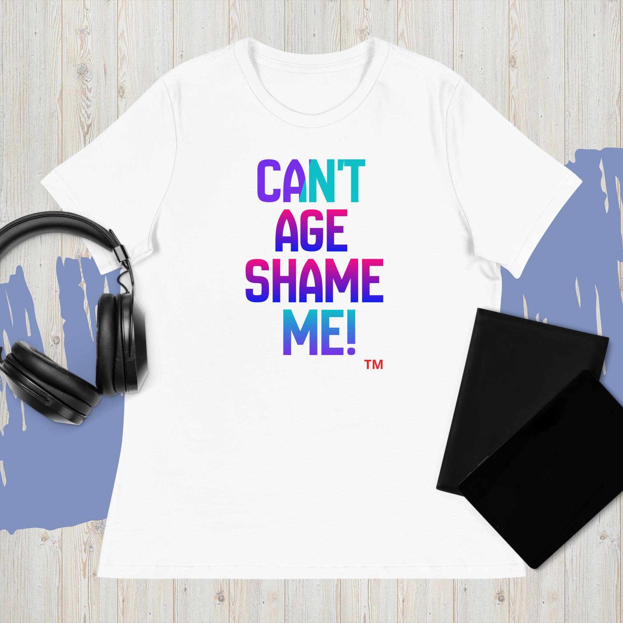 White t-shirt with colorful text 'CAN'T AGE SHAME ME!' on a wooden surface with headphones and a black mat.