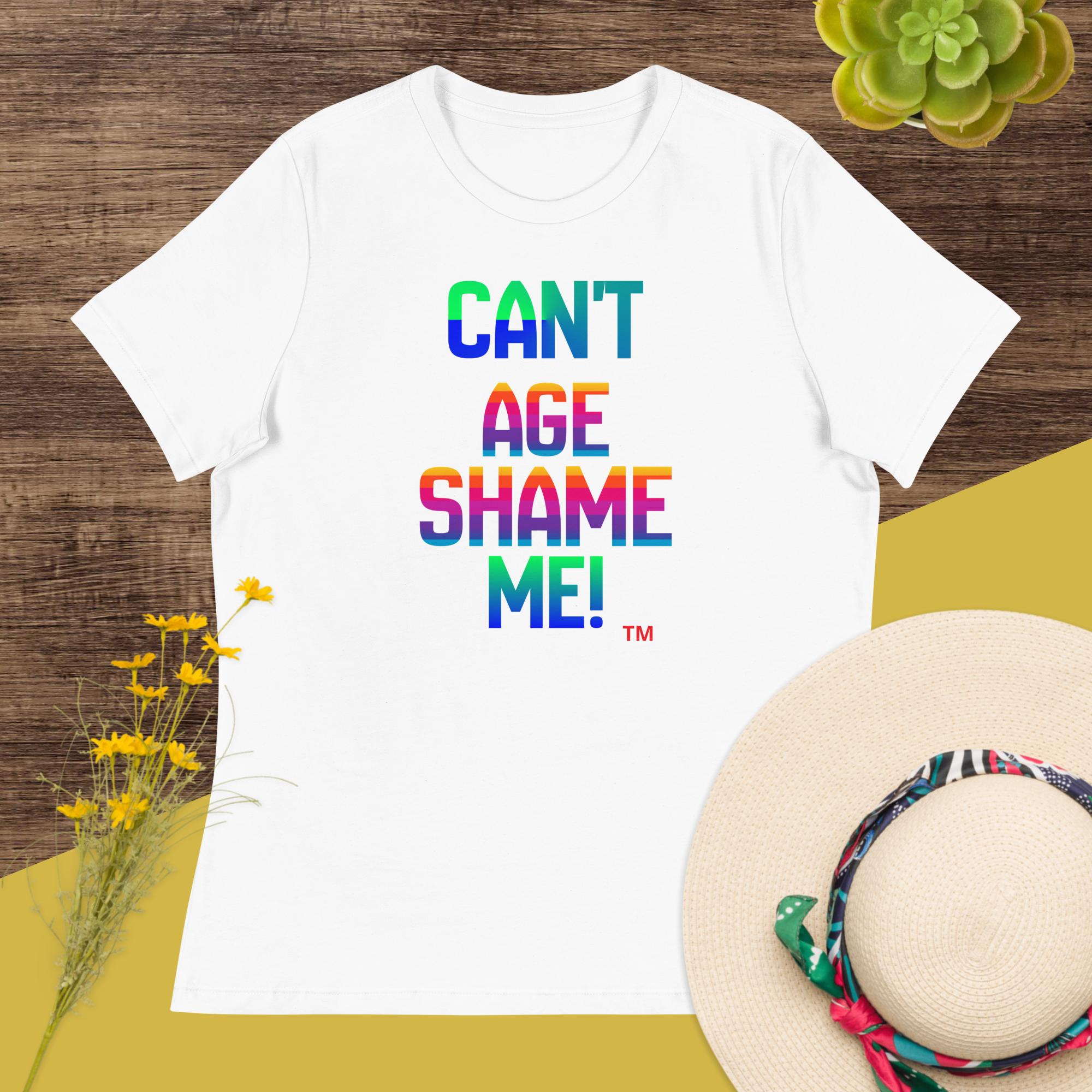 White t-shirt with colorful text 'CAN'T AGE SHAME ME!' on a wooden surface with flowers and a hat.