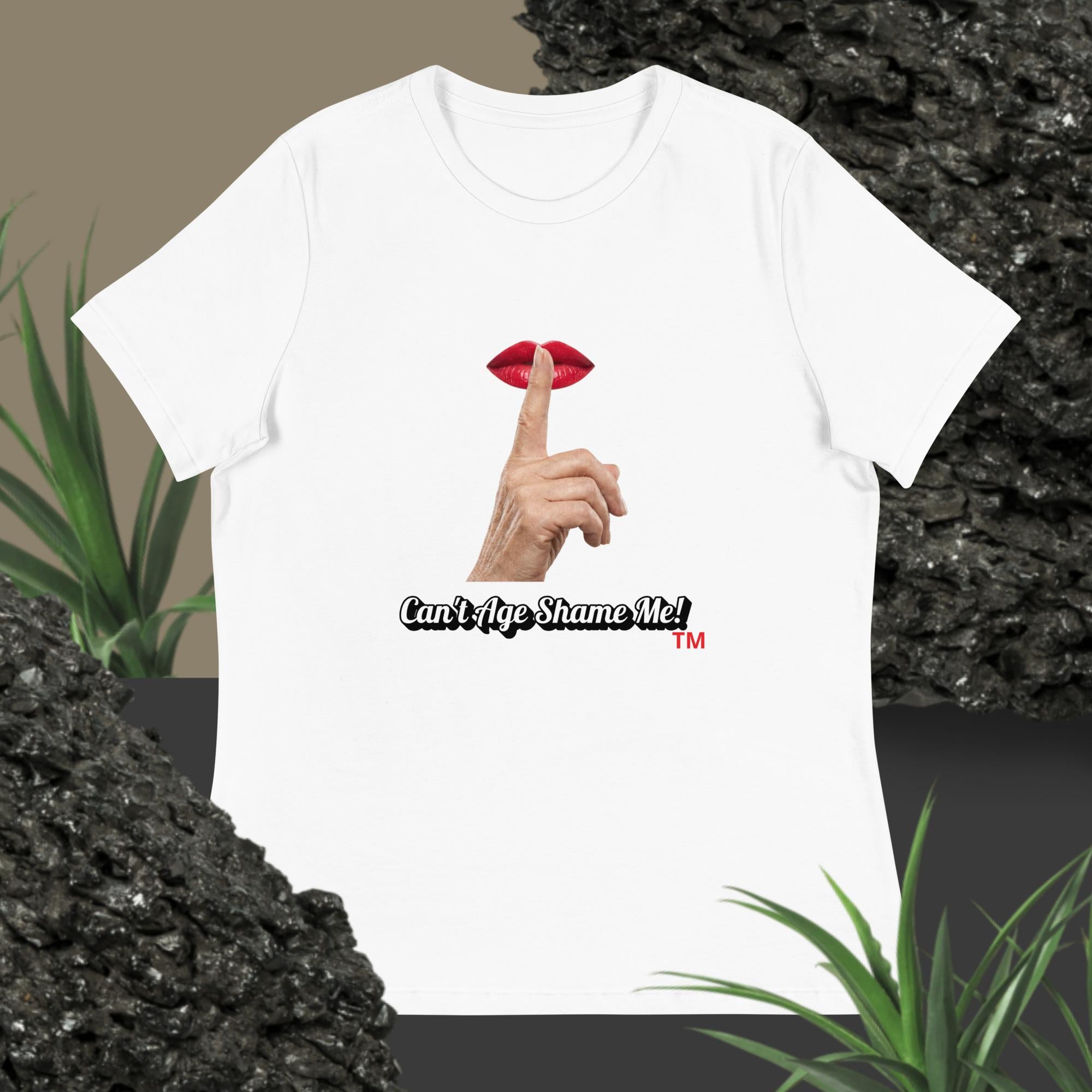 White t-shirt with graphic design of finger on mouth and text 'Can't Age Shame Me' on a textured surface with plants.