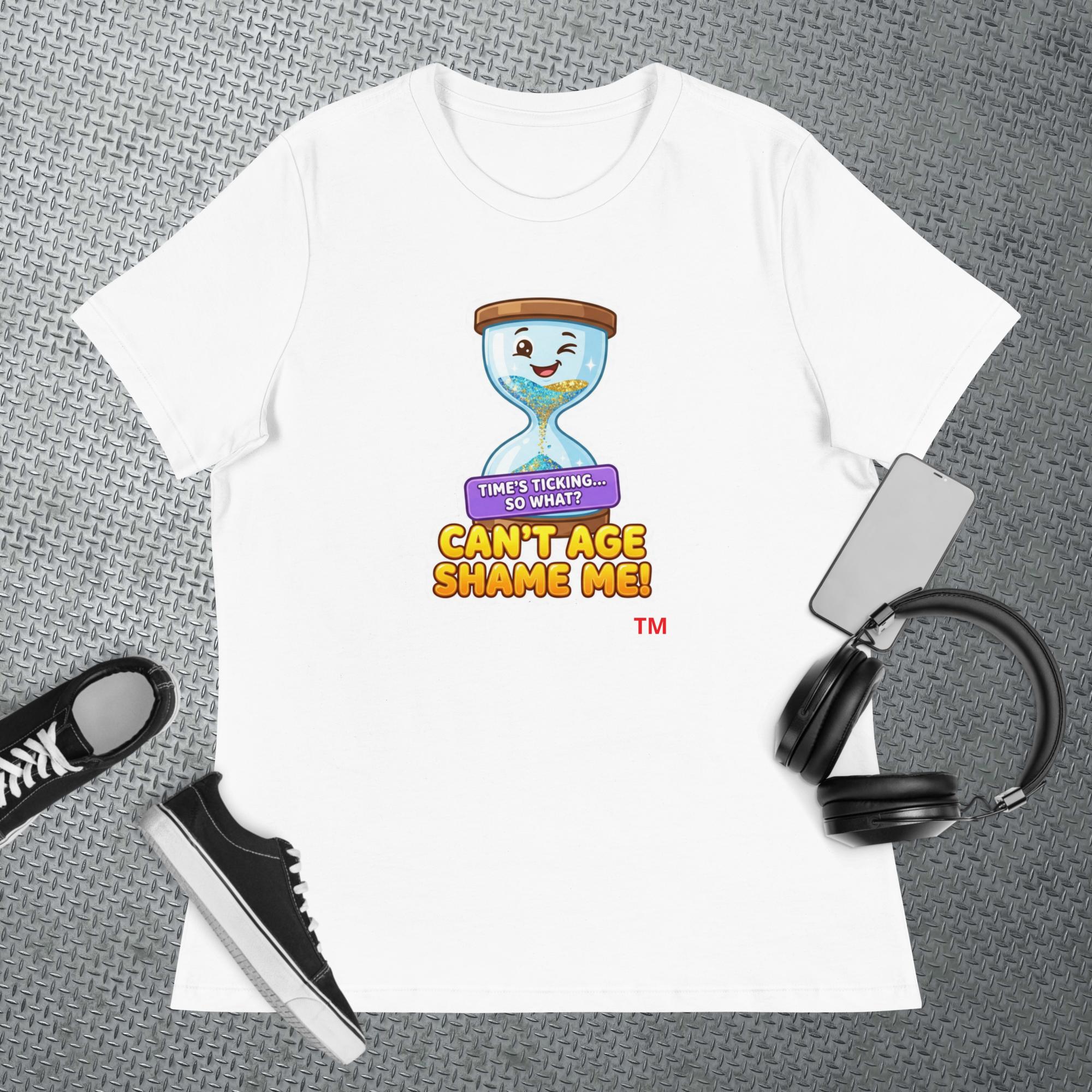 White t-shirt with a cartoon hourglass graphic design and text 'Can't Age Shame Me' graphic on a textured gray surface with a sneaker and headphones