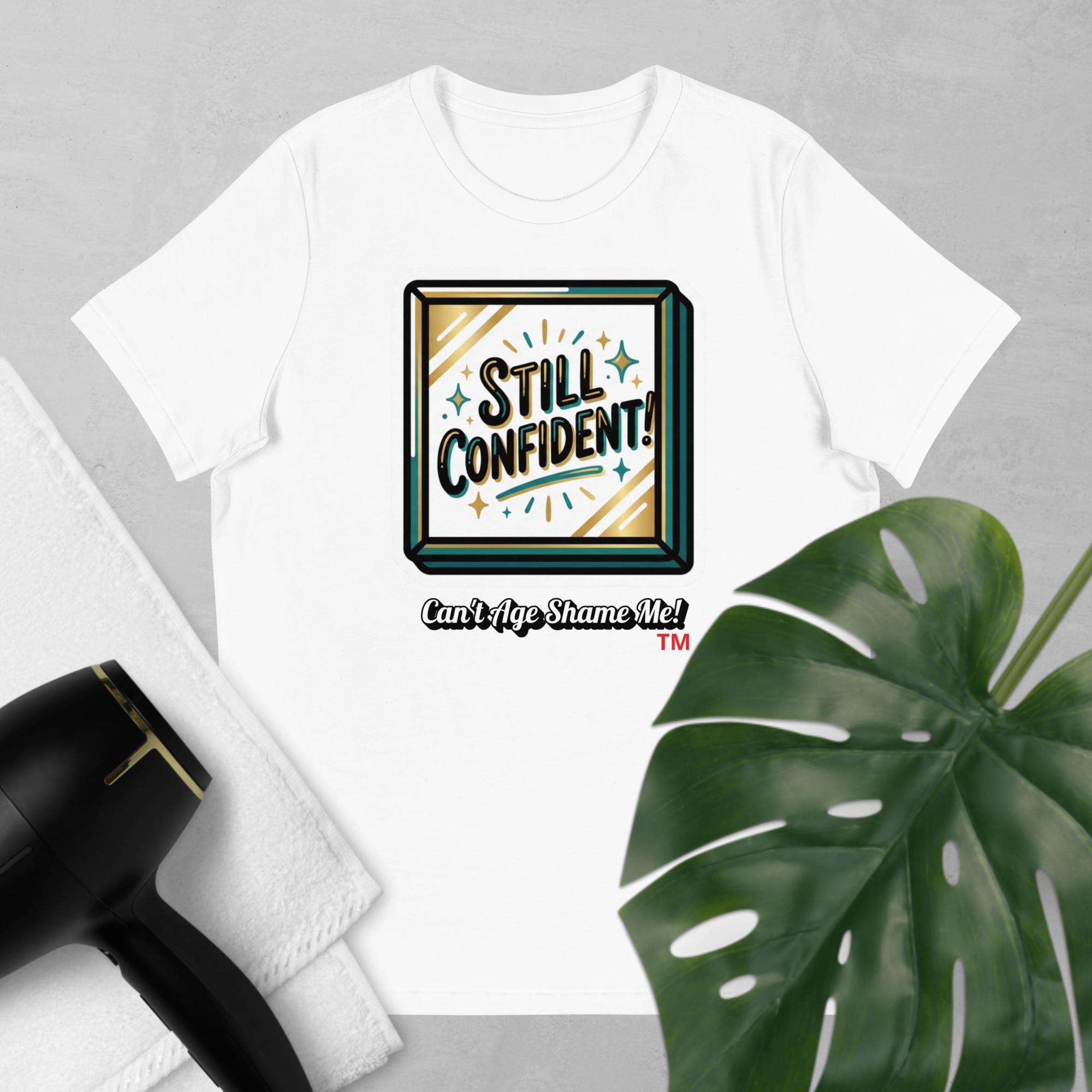 White t-shirt with 'Still Confident! Can't Age Shame Me!' graphic on a gray background with a plant and hair dryer.